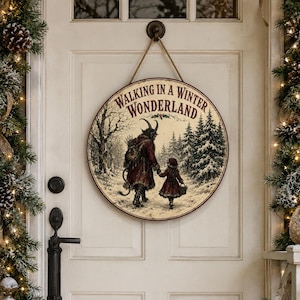 Könnte beinhalten: Ein rundes Holzschild, das an einem Seil hängt und eine Illustration einer gehörnten Figur in einem roten Mantel zeigt, die mit einem Kind durch einen verschneiten Wald geht. Der Text WALKING IN A WINTER WONDERLAND steht oben in Dunkelrot.