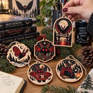 May include: Five wooden ornaments featuring Mothman illustrations. One is arched, one is hexagonal, and three are circular. The designs include a moth flying over a bridge, a moth in a forest, a moth holding a mug, and a moth by a campfire.