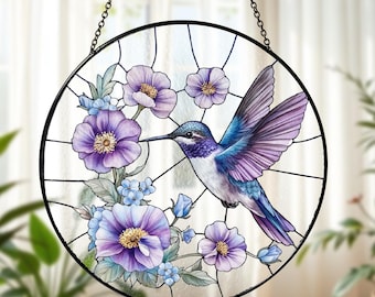 Op de afbeelding: Een rond gebrandschilderd glazen raam met een kolibrie in vlucht, omringd door paarse bloemen en blauwe accenten. De vogel heeft blauwe en paarse veren. Het raam is zwart omlijst en hangt aan een ketting.