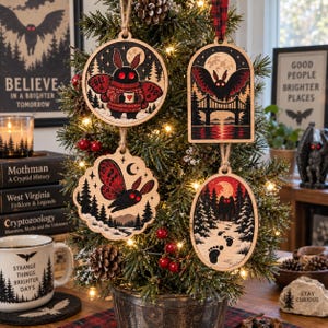 May include: Four wooden Christmas ornaments featuring Mothman illustrations in red, black, and cream. The designs include a moth holding a mug, a moth flying over a bridge, a moth in a forest, and a silhouette walking through snowy woods.