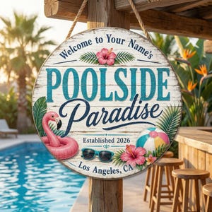 May include: A circular wooden sign with the text "Welcome to Your Name's POOLSIDE Paradise" in teal and navy blue. It features a pink flamingo float, sunglasses, and a beach ball. The sign also says "Established 2026" and "Los Angeles, CA".