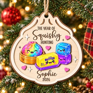 May include: A wooden ornament featuring the text THE YEAR OF SQUISHY HUNTING, Sophie, and 2026. The design includes a rainbow dumpling in a steamer, a yellow stick of butter labeled 4 oz., and two stacked blue and purple cubes with faces.