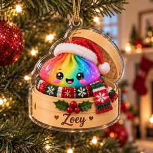 May include: A clear acrylic Christmas ornament featuring a rainbow-colored dumpling character wearing a red Santa hat and a red, green, and white snowflake-patterned scarf. The dumpling sits in a bamboo steamer basket labeled with the name Zoey.