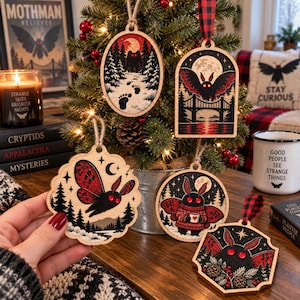 May include: A collection of five wooden ornaments featuring Mothman illustrations in red, black, and white. The designs include a flying creature, a figure in a snowy forest, a moth over a bridge, a moth holding a mug, and a moth with pine branches.