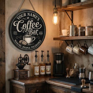 May include: A round black and white coffee bar sign with the text "YOUR NAME'S Coffee Bar" and the phrase "MAKING PEOPLE TOLERABLE". The sign features a coffee cup and cookies. The sign is hanging on a wall.