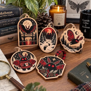 May include: Five wooden ornaments featuring Mothman illustrations with red eyes. Designs include a moth over a bridge, a forest path with footprints, a moth in flight, a moth holding a mug, and a moth with pinecones. Each has a hanging loop.