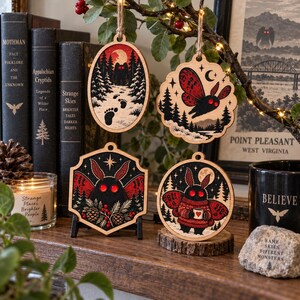 May include: Four wooden ornaments featuring Mothman illustrations. One oval shows footprints in snow, one scalloped circle shows a moth in flight, one hexagon shows a moth with pinecones, and one circle shows a moth holding a mug, all with red accents.