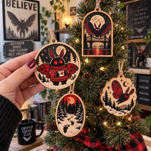 May include: Four wooden ornaments featuring Mothman illustrations. One circular ornament shows a Mothman in a red sweater holding a mug. Another is arched with a Mothman flying over a bridge. An oval one shows footprints in snow, and a scalloped one shows a flying Mothman.