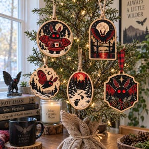 May include: Five wooden ornaments featuring Mothman illustrations in black, red, and white. Designs include a moth in a sweater, a moth flying over a bridge, a moth in flight, a moth in a snowy forest with footprints, and a moth with pinecones.