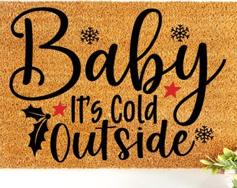 May include: A rectangular coir doormat with the phrase "Baby It's Cold Outside" in black script. The design includes snowflakes, a red star, and holly. A small potted plant with green leaves is in the lower right corner.