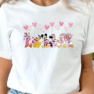 May include: White t-shirt featuring a colorful graphic of Disney characters, including Mickey and Minnie Mouse, holding heart-shaped balloons. The characters are in pink, black, and yellow. The shirt is made of a soft material.