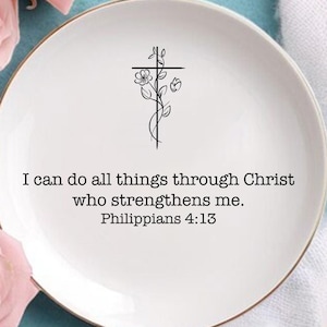May include: A white ceramic plate with a gold rim, featuring a black floral cross design and the text "I can do all things through Christ who strengthens me. Philippians 4:13." Pink roses and a white cloth are arranged around the plate.