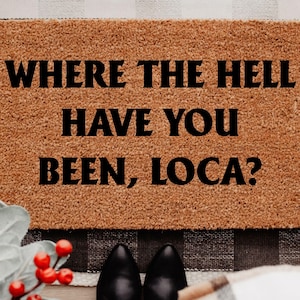 May include: A rectangular doormat with the text "WHERE THE HELL HAVE YOU BEEN, LOCA?" in black letters. The mat is brown and sits on a black and white checkered rug. Black boots and a plaid blanket are visible.