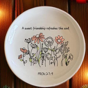 May include: White ceramic plate with a floral design in black, pink, and green. The plate features the text "A sweet friendship refreshes the soul" and "PROV.27:9". The plate is surrounded by Christmas decorations.