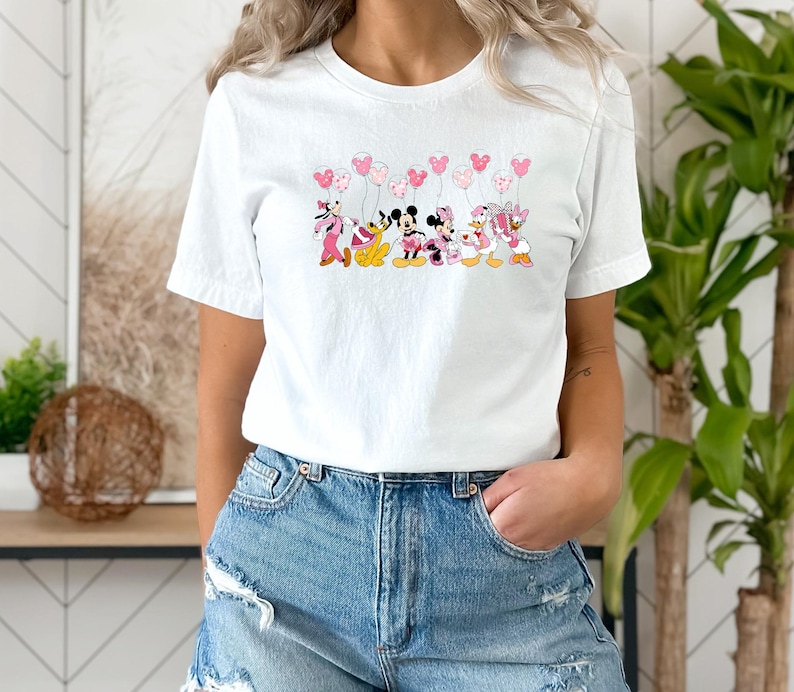 May include: White t-shirt featuring a colorful graphic of Disney characters, including Mickey and Minnie Mouse, holding heart-shaped balloons. The characters are in pink, black, and yellow. The shirt is made of a soft material.