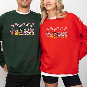 May include: Green and red crewneck sweatshirts featuring a cartoon design of Disney characters. The design includes Mickey Mouse, Minnie Mouse, Donald Duck, and Daisy Duck. Pink heart shapes are above the characters. The sweatshirts are worn by two people.