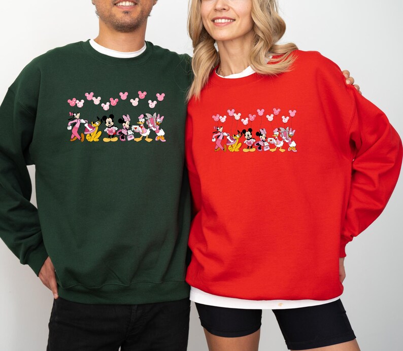 May include: Green and red crewneck sweatshirts featuring a cartoon design of Disney characters. The design includes Mickey Mouse, Minnie Mouse, Donald Duck, and Daisy Duck. Pink heart shapes are above the characters. The sweatshirts are worn by two people.