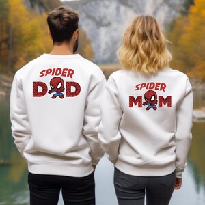 Puede incluir: Sudaderas blancas con el texto "SPIDER DAD" y "SPIDER MOM" en rojo, con un gr&aacute;fico de Spider-Man. Las sudaderas las llevan dos personas de pie frente a un lago y &aacute;rboles.