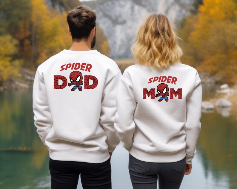 Puede incluir: Sudaderas blancas con el texto "SPIDER DAD" y "SPIDER MOM" en rojo, con un gr&aacute;fico de Spider-Man. Las sudaderas las llevan dos personas de pie frente a un lago y &aacute;rboles.