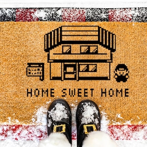 May include: A rectangular doormat with a pixelated house design and the words "HOME SWEET HOME" in black. The mat is tan with a red and black plaid border. Black shoes with gold buckles are visible.