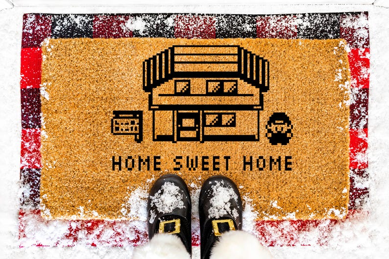 May include: A rectangular doormat with a pixelated house design and the words "HOME SWEET HOME" in black. The mat is tan with a red and black plaid border. Black shoes with gold buckles are visible.