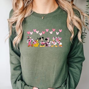 May include: An olive green sweatshirt featuring a line of cartoon characters including Mickey and Minnie Mouse, Pluto, and Daisy and Donald Duck. Pink heart-shaped mouse ears are above the characters. The sweatshirt is long-sleeved.