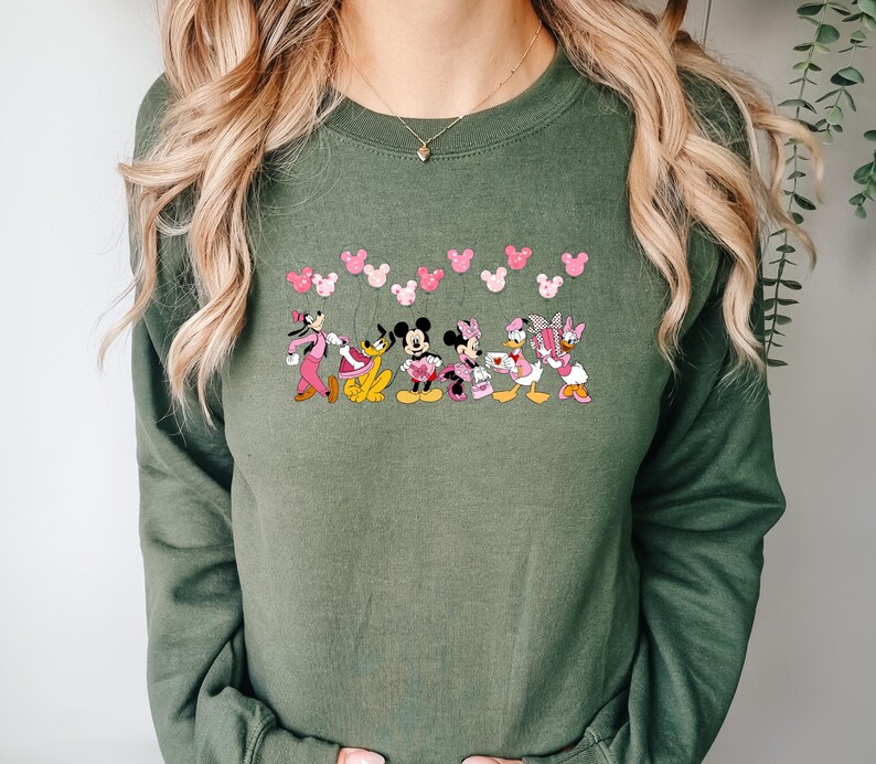 May include: An olive green sweatshirt featuring a line of cartoon characters including Mickey and Minnie Mouse, Pluto, and Daisy and Donald Duck. Pink heart-shaped mouse ears are above the characters. The sweatshirt is long-sleeved.