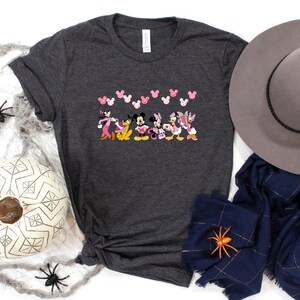 May include: Dark gray t-shirt featuring a colorful graphic of Disney characters, including Mickey and Minnie Mouse, and Daisy and Donald Duck. Pink Mickey Mouse head shapes are above the characters. A gray hat and navy blue scarf are next to the shirt.