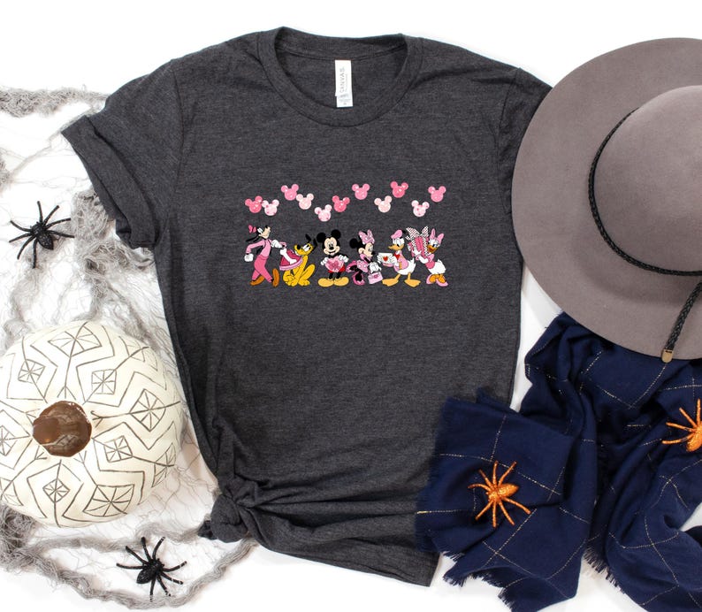 May include: Dark gray t-shirt featuring a colorful graphic of Disney characters, including Mickey and Minnie Mouse, and Daisy and Donald Duck. Pink Mickey Mouse head shapes are above the characters. A gray hat and navy blue scarf are next to the shirt.
