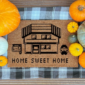 May include: A brown doormat with a pixelated house design and the words "HOME SWEET HOME" in black. The mat is surrounded by various pumpkins in shades of orange, yellow, and green, creating a fall theme.
