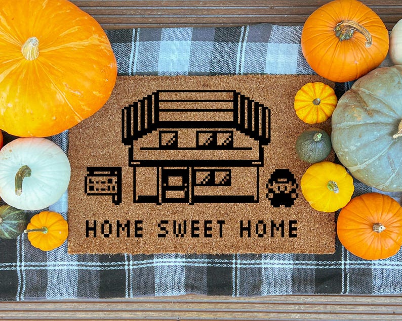 May include: A brown doormat with a pixelated house design and the words "HOME SWEET HOME" in black. The mat is surrounded by various pumpkins in shades of orange, yellow, and green, creating a fall theme.