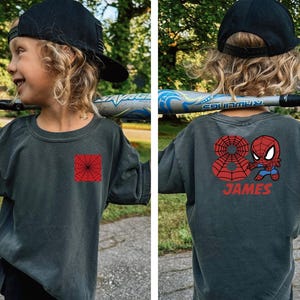 Puede incluir: Camiseta gris oscuro con un dise&ntilde;o de telara&ntilde;a roja en la parte delantera y un gr&aacute;fico de Spider-Man en la parte trasera. La parte trasera de la camiseta tambi&eacute;n presenta el nombre "JAMES" en rojo. La camiseta la lleva un ni&ntilde;o.