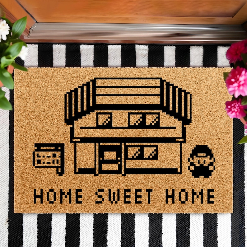 May include: A rectangular doormat with a pixelated house design in black on a tan background. The words "HOME SWEET HOME" are printed in black block letters below the house graphic. The mat is placed on a black and white striped surface.