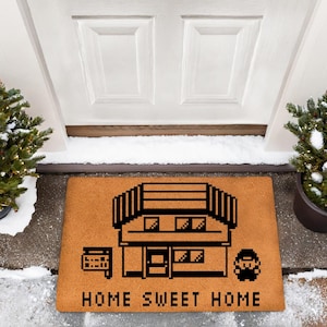 May include: A rectangular doormat with a pixelated house design and the words "HOME SWEET HOME" in black. The mat is brown and sits in front of a white door, flanked by potted evergreen trees.