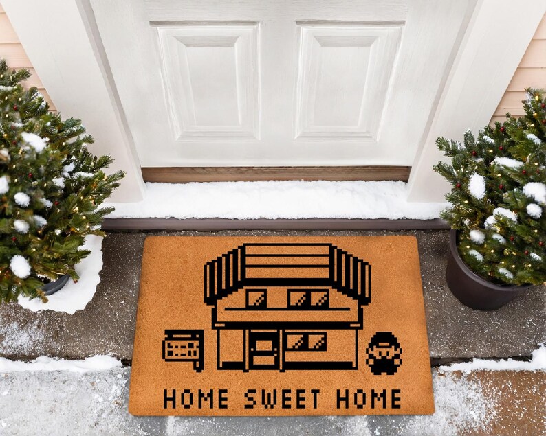 May include: A rectangular doormat with a pixelated house design and the words "HOME SWEET HOME" in black. The mat is brown and sits in front of a white door, flanked by potted evergreen trees.
