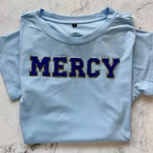 Puede incluir: Una camiseta azul claro con la palabra "MERCY" en letras azules y doradas.
