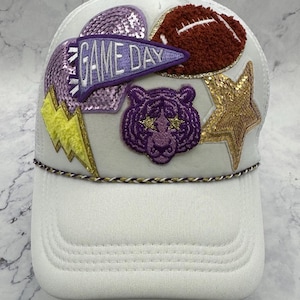 Op de afbeelding: Witte truckercap met verschillende kleurrijke patches, waaronder een paars hart met pailletten, een paars-witte vlag met de tekst "GAME DAY", een bruine voetbal, een paarse tijger en een gouden ster.