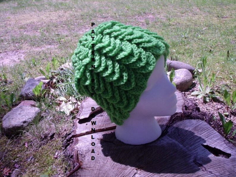 May include: A bright green, crocheted hat with a textured, layered design. The hat is displayed on a white mannequin head, set against a natural outdoor backdrop. The text "PATTERN PDF DOWNLOAD" is vertically printed on the left side of the hat.