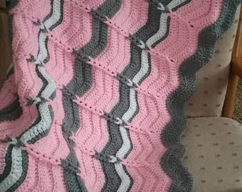 May include: A handmade crochet blanket with a wavy design. The blanket has stripes of pink, grey, and white. The edges are finished with a scalloped border. Small white accents are evenly spaced along the grey stripes.