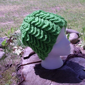 May include: A bright green, crocheted hat with a textured, layered design. The hat is displayed on a white mannequin head, set against a natural outdoor backdrop of grass, rocks, and a tree stump.