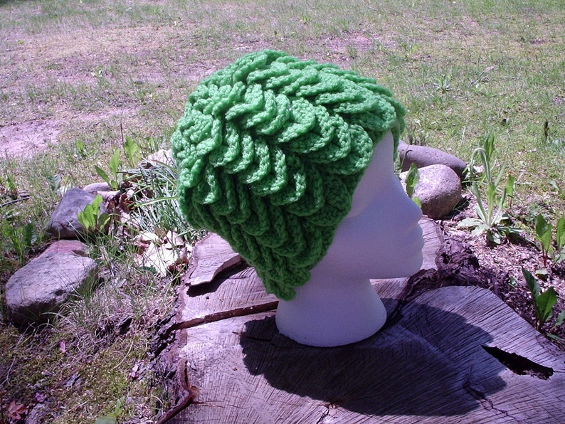 May include: A bright green, crocheted hat with a textured, layered design. The hat is displayed on a white mannequin head, set against a natural outdoor backdrop of grass, rocks, and a tree stump.