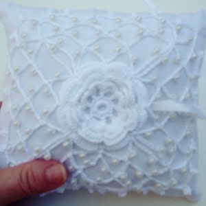May include: White crocheted ring pillow with a pearl trim and a large white crocheted flower in the center.