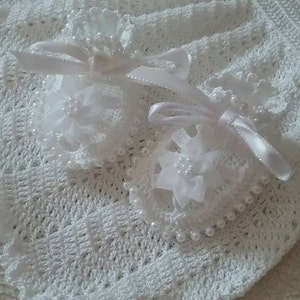 May include: A white crocheted baby blanket with a scalloped edge. Two white floral hair accessories with pearl accents and satin ribbons are placed on the blanket. The blanket and accessories are made of soft, delicate materials.