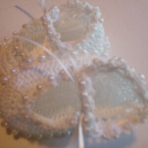 May include: A pair of white, crocheted baby booties. The booties are adorned with small, pearl-like beads around the edges and tied together with a white ribbon. The delicate design suggests a handmade item.