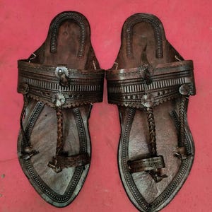May include: A pair of handcrafted leather sandals in a dark brown hue. The sandals feature intricate embossed detailing, a braided toe strap, and a decorative buckle. The open-toe design and flat sole suggest a comfortable, casual style.