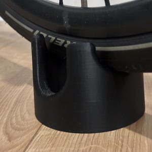 May include: Black bicycle tire resting on a black 3D-printed stand. The tire has the word "PIRELLI" printed on it. The stand is cylindrical with a curved top to cradle the tire.