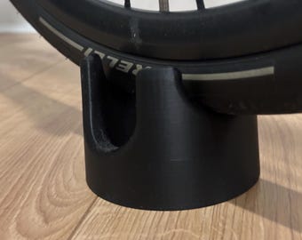 May include: Black bicycle tire resting on a black 3D-printed stand. The tire has the word "PIRELLI" printed on it. The stand is cylindrical with a curved top to cradle the tire.