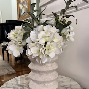 May include: A white ceramic vase with a textured, ridged design holds a bouquet of white hydrangeas and olive branches. The vase sits on a marble-topped table, creating a neutral and elegant floral arrangement.