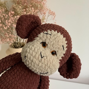Può includere: Un peluche a forma di scimmia realizzato in filato testurizzato marrone scuro e beige. La scimmia presenta orecchie rotonde, un viso beige chiaro e due occhi in plastica color ambra. Dettagli in filo nero ricamato definiscono sopracciglia e naso.