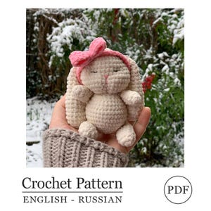 May include: A crocheted bunny toy in cream color with a pink bow headband. The bunny is held in a hand against a snowy background. The text "Crochet Pattern" and "ENGLISH - RUSSIAN" is at the bottom of the image.